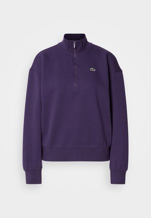 Vijoličen flis pulover s polzadrgo, rebrastimi manšetami in pasom. Na prsih je majhen zeleni krokodilov logotip.