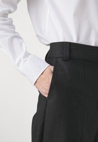 Čierne pruhované nohavice s klasickým strihom, obsahujúce opasok a bočnú kapsu, v kombinácii s bielou košeľou s dlhým rukávom a gombíkmi.
