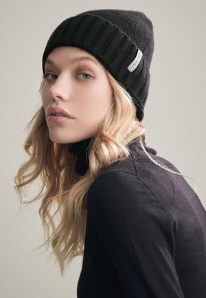 Zwarte geribbelde gebreide beanie met een label aan de zijkant, gedragen met een zwarte coltrui. Haar gestyled in losse golven. Neutrale achtergrond.