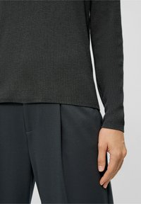 Eine Person trägt einen dunklen, strukturierten Strickpullover und dunkle, plissierte Hosen, mit der rechten Hand entspannt an ihrer Seite.