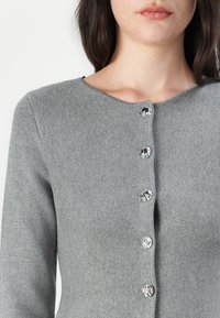 Femme portant un cardigan en maille gris avec cinq boutons ronds argentés brillants, visible des épaules jusqu'au milieu du torse.