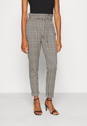 Kobieta ubrana w wysokie spodnie w kratę z przewiązanym paskiem oraz czarne sandały na obcasie z paskami, stoi na tle białego tła.