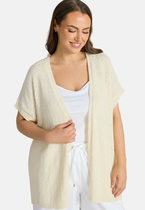 Uśmiechnięta kobieta w kremowym, krótkim kardiganie na otwartym przodzie, nosząca pod nim białą koszulkę na ramiączkach oraz białe spodnie z regulowanym sznurkiem.