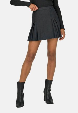 Woman wearing a dark pleated mini skirt, black long-sleeve top, and black heeled ankle boots, standing against a plain background.