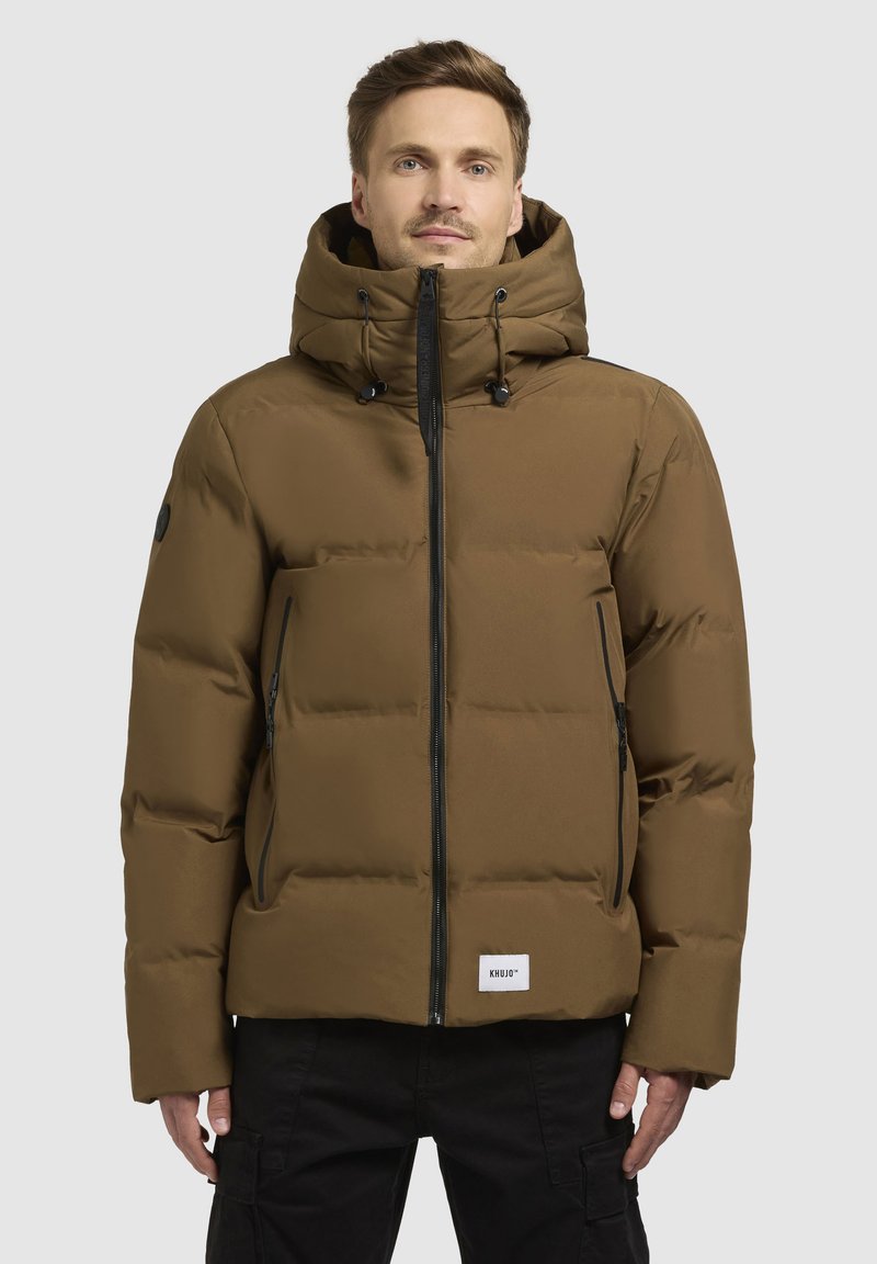 Giubbotto puffo marrone con cappuccio, chiusura con zip e tasche laterali. Presenta accessori neri e un cordino regolabile sul cappuccio.