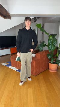 Junger Mann trägt schwarzes Langarmshirt, beige Hose und Turnschuhe und steht in einem modernen Wohnzimmer mit einem braunen Sofa und einer großen grünen Pflanze.