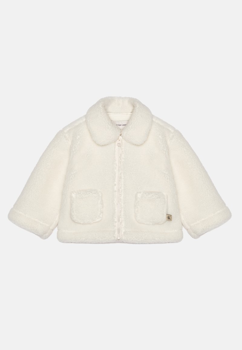 Bílá fleecová bunda s límcem na knoflíky, se zipem v přední části a dvěma předními kapsami. Má jemnou texturu a malou etiketu s logem na lemu.