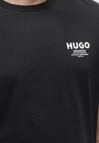 Μαύρο T-shirt με στρογγυλή λαιμόκοψη, λευκό λογότυπο "HUGO" και μικρό κείμενο στο αριστερό στήθος, φορεμένο από άτομο ορατό από τον λαιμό μέχρι το μέσο του κορμού.