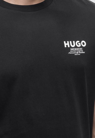 Черен тениска с кръгъл дизайн и бял логотип "HUGO" и малък надпис от лявата страна на гърдите, носена от човек, видим от врата до средата на тялото.