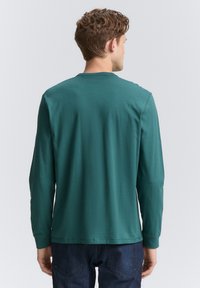 Langärmliges T-Shirt in einem tiefen Teal-Farbton, aus weicher Baumwolle gefertigt, mit Rundhalsausschnitt und geradem Saum, von hinten gezeigt.