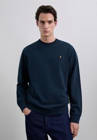 Sweat-shirt bleu marine en tissu doux avec un col rond. Présente un petit emblème jaune sur la poitrine ainsi que des poignets et un ourlet côtelés.