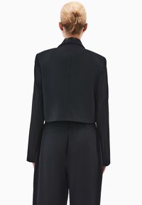 Blazer nero cropped con maniche lunghe, texture liscia e dettagli sul collarino. La vista posteriore mostra una vestibilità su misura e linee pulite.