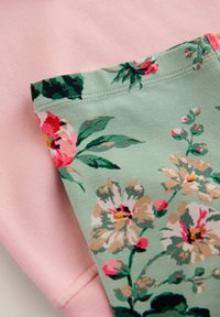 Bombažna oblačila, ki vključujejo mehak roza material, v kombinaciji z zelenim panelom, okrašenim z velikimi cvetličnimi vzorci v roza, krem in zeleni barvi.