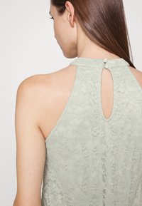Top halter em renda verde menta com costas em forma de chave e fecho de botão. O tecido tem um padrão floral, destacando sua textura delicada.