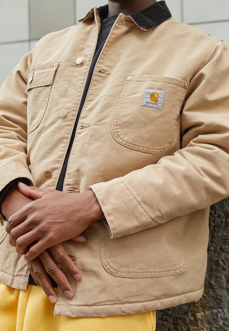 Brun canvasjakke med sort krave, featuring guld hardware, forlommer og et Carhartt logo patch på brystlommen.