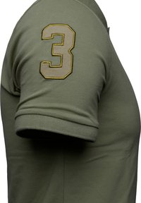 Oliivinvihreä polo-paita lyhyillä hihoilla, jossa on edessä näkyvä brodattu numero 3 vaaleassa beessissä ja keltaisessa vasemmalla olkapäällä.