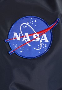 Blauw NASA-embleem met witte letters, rode vector-swoosh, witte baan en sterren op een donkere stoffen achtergrond.