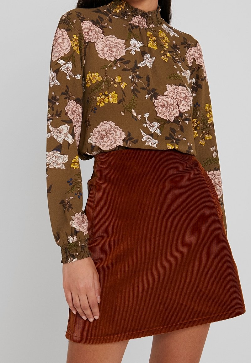 Brun blus med blommönster och långa ärmar, med rynkad halsringning; ihop med en rostfärgad manchesterkjol med vertikal ribbad struktur.