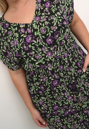 Femme portant une robe noire à motif floral violet et vert, encolure carrée et manches courtes, main posée sur la hanche.