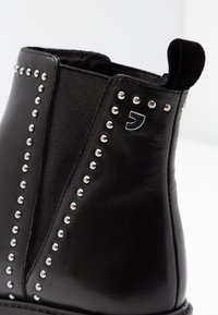 Botín negro de cuero con tachuelas plateadas a lo largo de las costuras y un panel lateral texturizado. Incluye una pequeña pestaña para facilitar el calzado.