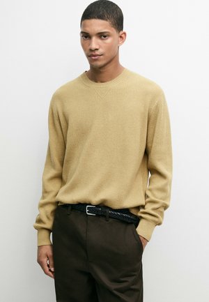 Jonge man die een beige geribde trui en donkere broek met een gevlochten zwarte riem draagt, staand tegen een effen witte achtergrond.