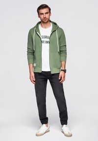 Grøn zip-up hættetrøje med hvide snore over en hvid T-shirt med sort tekst. Parret med mørke jeans og hvide sneakers.