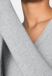 Gros plan sur le cou et le haut de la poitrine d'une personne portant un pull en tricot gris clair avec un col en V.