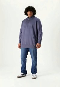 Sudadera en azul apagado con cuello de cremallera de un cuarto, bolsillo delantero y texto en relieve. Combinada con vaqueros azules y zapatillas blancas.