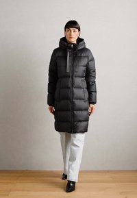 Abrigo puffer negro largo con capucha, que cuenta con un cierre de cremallera, múltiples secciones acolchadas horizontales y un corte holgado. Textura suave y diseño elegante.
