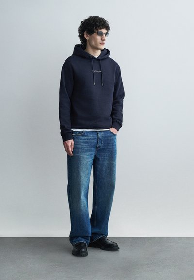 Homme aux cheveux bouclés portant des lunettes de soleil noires, un sweat à capuche marine, un jean bleu et des chaussures noires, debout devant un fond gris clair uni.