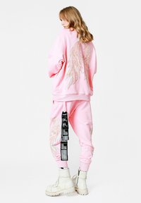 Conjunto de sudadera y pantalones de chándal rosa con alas de ángel bordadas. El material parece suave; tiene un detalle de etiqueta en los pantalones. Botas blancas de estilo grueso.
