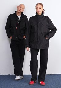 Two models wear black outer jackets with multiple pockets. One model pairs it with black pants and white shoes, while the other wears red shoes.
