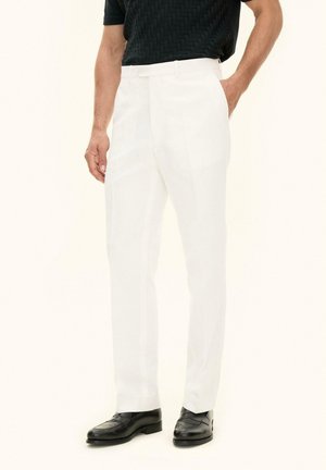 Mężczyzna w białych dopasowanych spodniach, czarnej teksturowanej koszuli z krótkim rękawem i czarnych skórzanych eleganckich butach, stojący z jedną ręką w kieszeni.