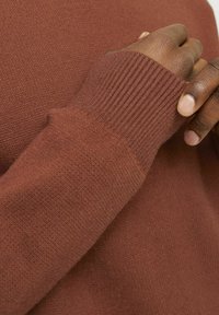 Primer plano de un suéter de punto marrón que muestra un puño acanalado y una textura suave, destacando su material blando y su color cálido.