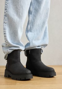 Botines negros de tobillo hechos de material suave, con suela gruesa, detalle de cremallera y punta redondeada, combinados con jeans ligeros de color azul claro arremangados.