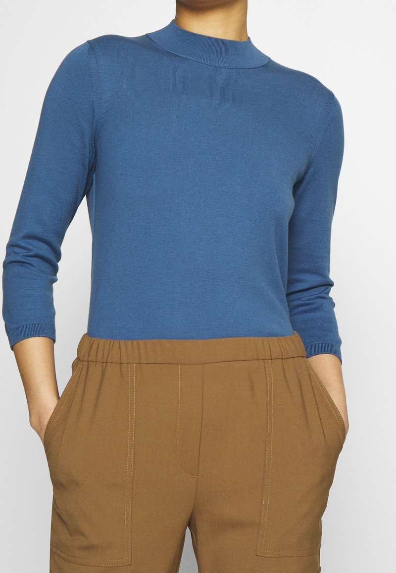 Niebieski sweter z prążkowanego materiału o wysokim dekolcie, w połączeniu z brązowymi spodniami z bocznymi kieszeniami i widocznymi detalami szwów.