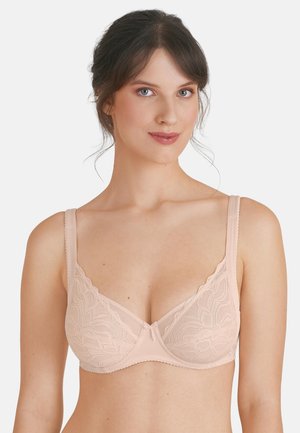 Frau mit dunklen Haaren trägt einen hellrosa Spitzen-BH mit Bügeln und blickt nach vorne vor einem schlichten weißen Hintergrund.