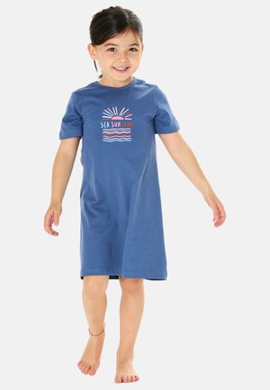 Smiling young girl walking barefoot wearing a blue dress with "Sea Sun Sand" and sun and wave graphics on the front.