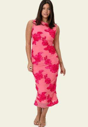 Mladá žena v bezrukávových ružových midi šatách s jasne ružovou kvetinovou výšivkou, stojaca uvoľnene na jednoduchom pozadí.