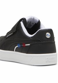 Zapatilla de cuero negra con suela blanca, que presenta acentos rojos y azules en el costado, y un logotipo redondo cerca del talón. Cierre de velcro.