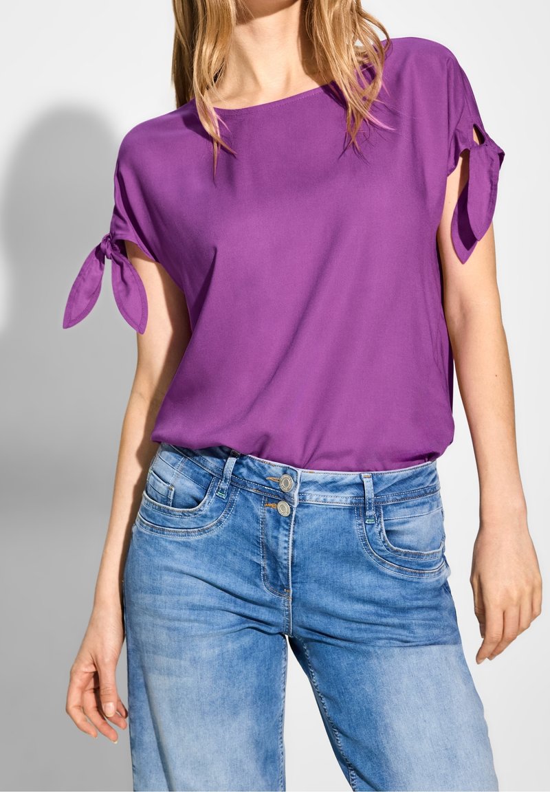 Haut violet à manches courtes avec des accents noués aux manches, associé à un jean en denim bleu clair avec une fermeture à double bouton et des poches.
