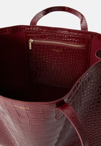 Bolsa tote em bordeaux profundo com textura de crocodilo, apresentando um bolso com fecho dentro e alças em couro liso. Interior forrado em tecido.