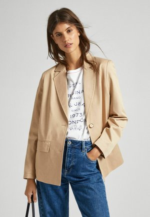 Blazer beige clair avec un bouton unique, deux poches avant et une coupe décontractée, porté sur un t-shirt blanc à lettres bleues et un jean foncé.