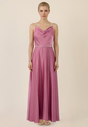 Frau steht vor einfachem Hintergrund und trägt ein bodenlanges, seidiges rosafarbenes Kleid mit dünnen Trägern und leichtem Wasserfallausschnitt.