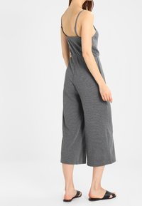 Mono de algodón gris con tirantes finos, una cintura ajustada y piernas anchas cortas. La tela tiene una textura suave. Combinado con sandalias negras tipo zueco.