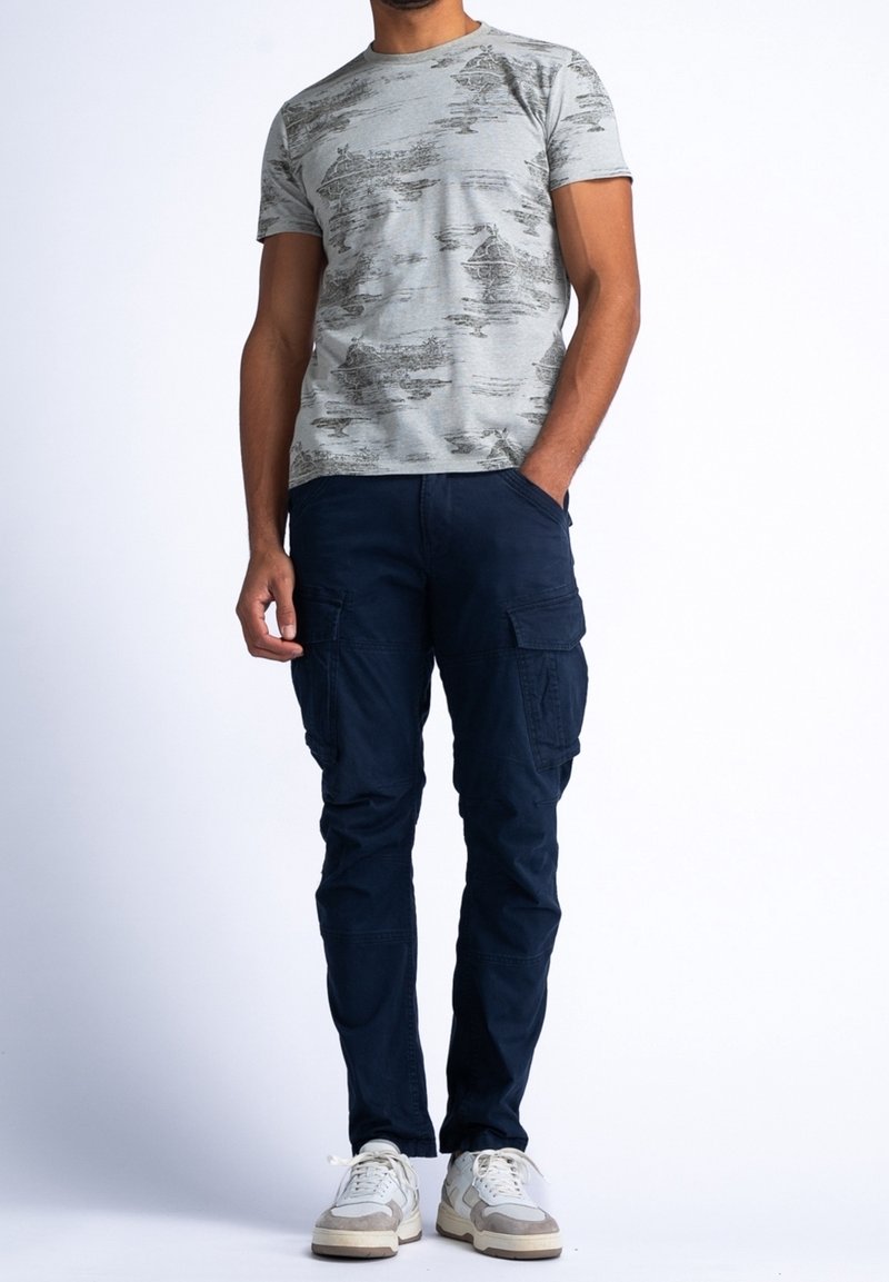 Homme portant un t-shirt à manches courtes gris clair à motifs, un pantalon cargo bleu foncé et des baskets blanches, debout avec une main dans la poche.