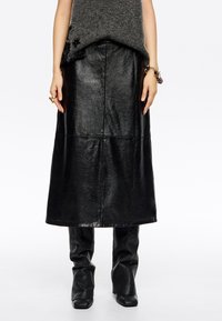 Falda midi de cuero negro con un acabado suave, presenta una silueta recta y costuras en panel. Llevada sobre pantalones ajustados negros.