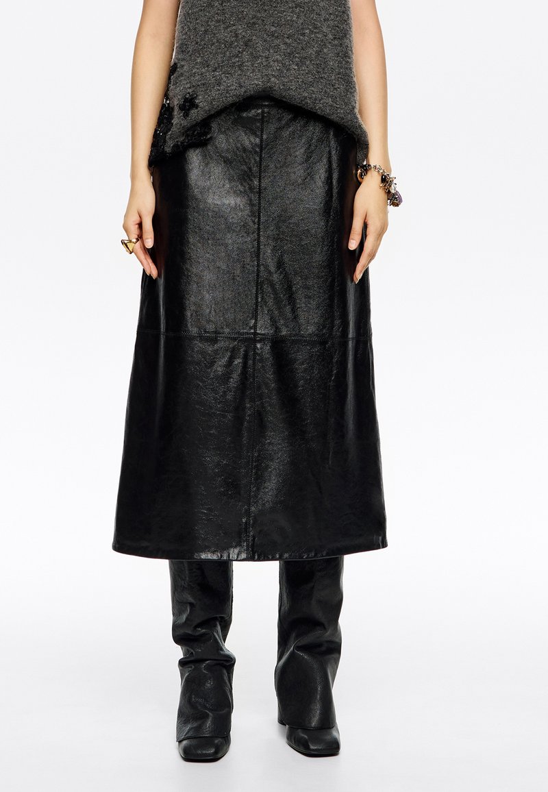 Falda midi de cuero negro con un acabado suave, presenta una silueta recta y costuras en panel. Llevada sobre pantalones ajustados negros.