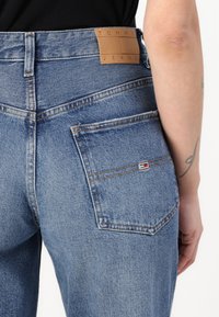 Korkeavyöti siniset farkut, joissa on takataskut, vaalea pesu, oranssi tikkaus ja ruskea nahkatee brändilogo vyötäröllä.