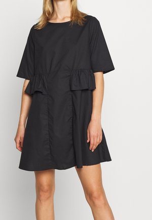 Kvinde iført en sort kortærmet minikjole med flæsedetaljer ved taljen mod en ensfarvet lys baggrund.